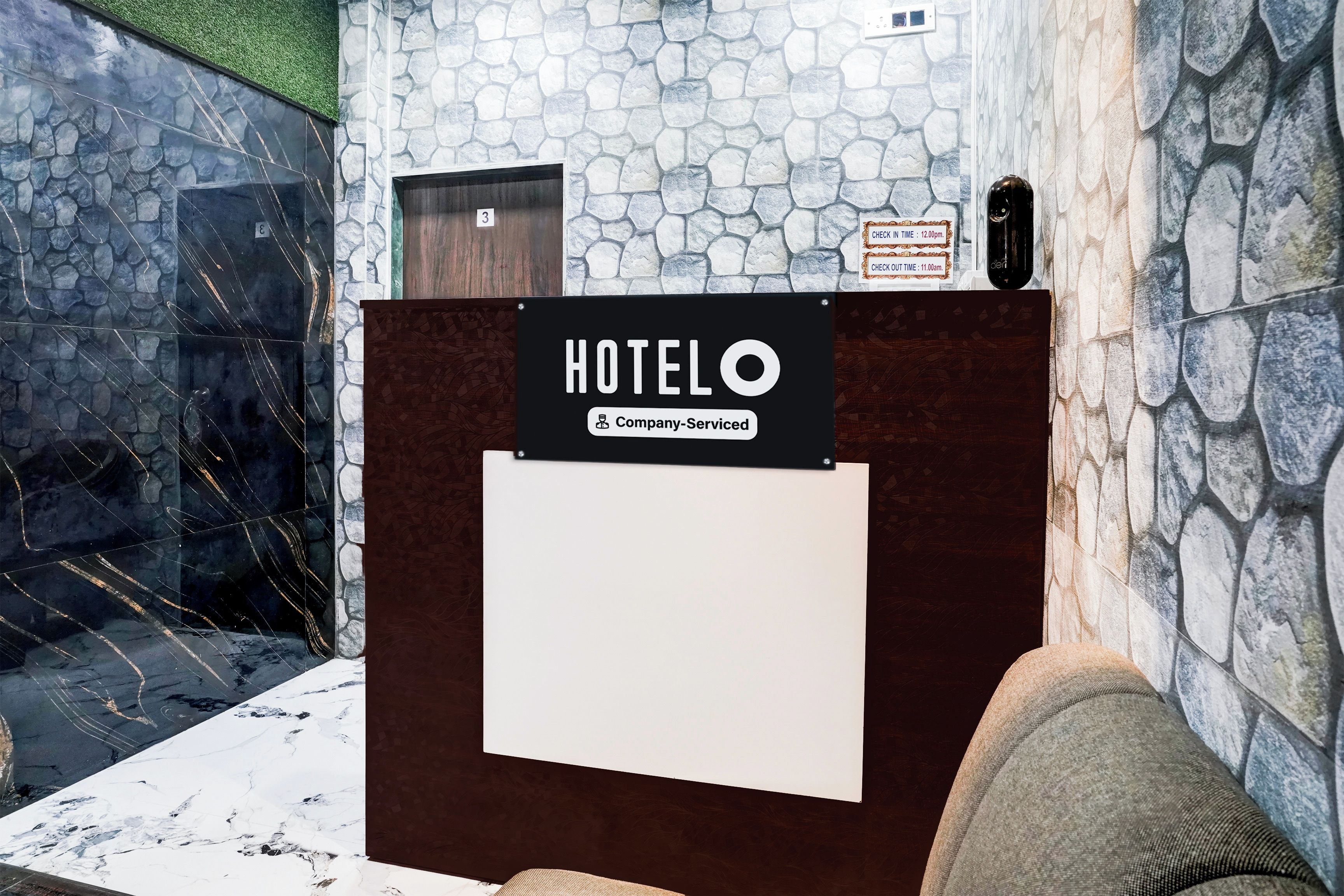 Una recepción de hotel con un fondo de pared de piedra, un cartel que indica habitaciones de servicio de la empresa y una pantalla con los horarios de registro de entrada y salida.