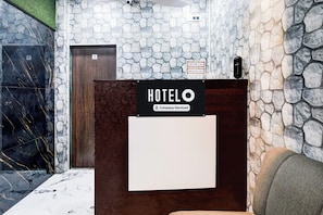 Un hall d’accueil d’hôtel avec un mur en pierre, un comptoir d’accueil en bois et un panneau indiquant les modalités d’arrivée et de départ.