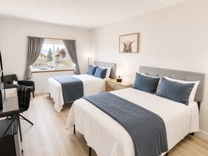 A hotel room with two beds, a desk, a chair, a window with curtains, and a framed picture on the wall.