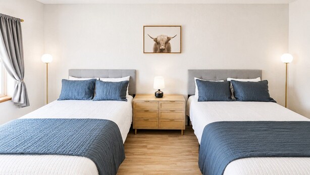 A hotel room with two beds, a bedside table with a lamp, a wooden floor, and a picture on the wall.
