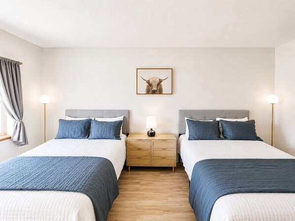 A hotel room with two beds, a bedside table with a lamp, a wooden floor, and a picture on the wall.