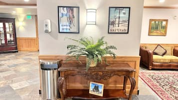 Un espace d’accueil avec un bureau en bois, une poubelle, une plante en pot, des tableaux encadrés au mur, un canapé et un tapis à motifs.