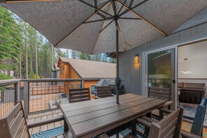 Una terraza de madera con una mesa y sillas, un espacio cubierto con una sombrilla, y una cabaña al fondo.