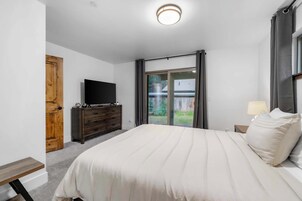 Un dormitorio con una cama, un televisor sobre un aparador de madera, una ventana con cortinas y una puerta.