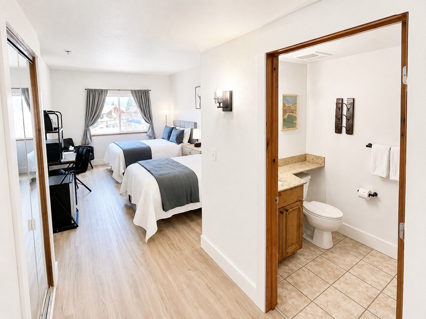 A hotel room with two beds, a desk, a chair, a window with curtains, a bathroom with a toilet and sink, and a door leading to another room.