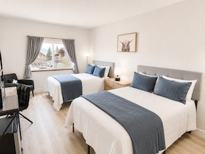 A hotel room with two beds, a desk, a chair, a window with curtains, and a framed picture on the wall.
