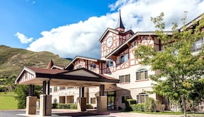 A traditional chalet-style hotel with a prominent clock tower, a covered entrance, and a mountainous backdrop.