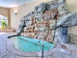 A swimming pool with a rocky wall backdrop, a metal ladder, and a tiled floor.