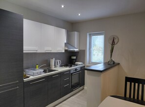 A modern kitchen with dark cabinetry, a gas stove, and a window with blinds. - Cozycorner (Konavle)