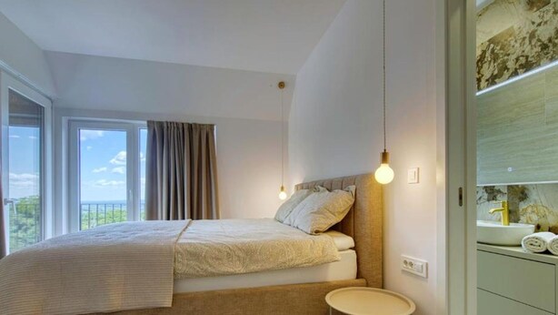 A bedroom with a bed, a bedside table, a window with curtains, and a view of the outdoors. - Villa One (Jastrebarsko)