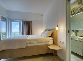 Un dormitorio con cama, mesita de noche, ventana con cortinas y vistas al exterior.