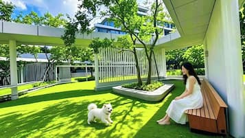 Una mujer sentada en un banco en un patio, acompañada por un perro, rodeada de follaje y arquitectura moderna.
