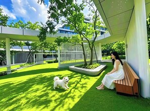 Eine Frau sitzt mit einem Hund auf einer Bank in einem Innenhof, umgeben von Grünflächen und moderner Architektur.