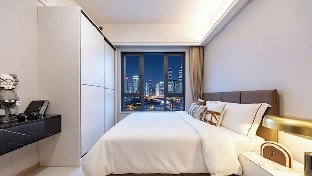A modern bedroom with a city view, a bed, bedside table, and a desk with a computer and headphones. - ruilantingyuan (shenzhen)