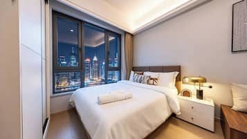 Una habitación de hotel moderna con un ventanal grande que ofrece vistas a la ciudad, una cama bien tendida, una mesita de noche con una lámpara y un pequeño aparador de madera.