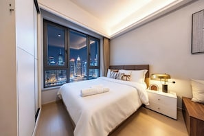 Ein modernes Hotelzimmer mit großem Fenster und Blick auf die Stadt, einem ordentlich bezogenen Bett, einem Nachttisch mit Lampe und einem kleinen Holzbeistelltisch.