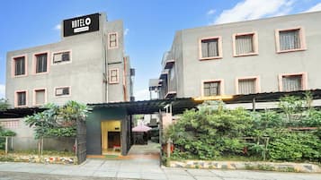 A building with a sign that reads 'HOTEL' and a company-serviced symbol. There are multiple windows with metal grilles, a covered entrance, and a small outdoor seating area with a table and chairs.