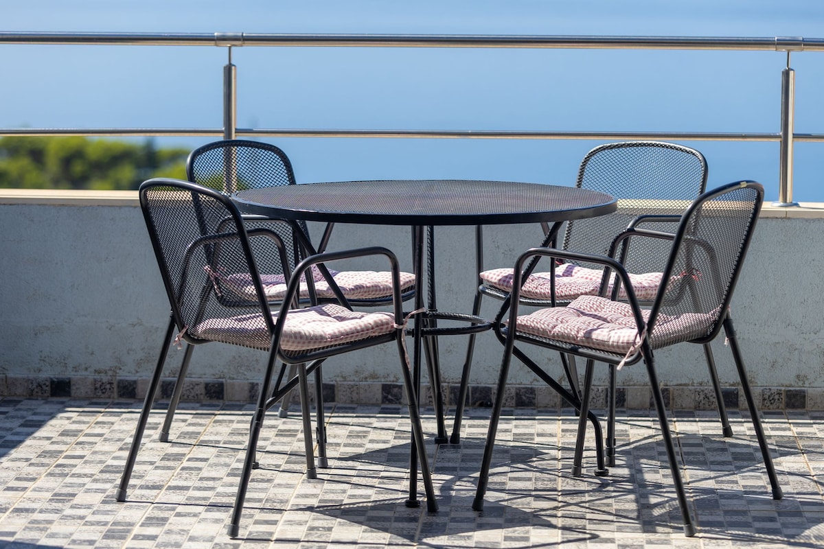 Ein runder Tisch mit vier Stühlen, jeweils mit gepolsterten Sitzflächen, auf einer gefliesten Terrasse mit Meerblick.
