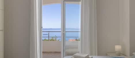 A bedroom with a bed, a nightstand, a lamp, and a view of the sea through a glass door.