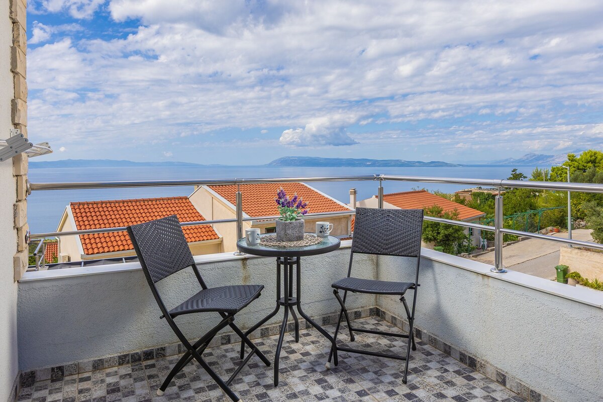 Ein Balkon mit zwei Korbstühlen und einem kleinen Tisch, mit Blick auf die Küste, mit roten Ziegeldächern und entfernten Bergen.