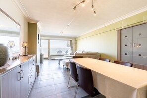 A modern kitchen with a long countertop, a dining area with a table and chairs, and a view of the outdoors through large windows. - Apartment in Zeedijk With Sea View Terrace (Blankenberge)