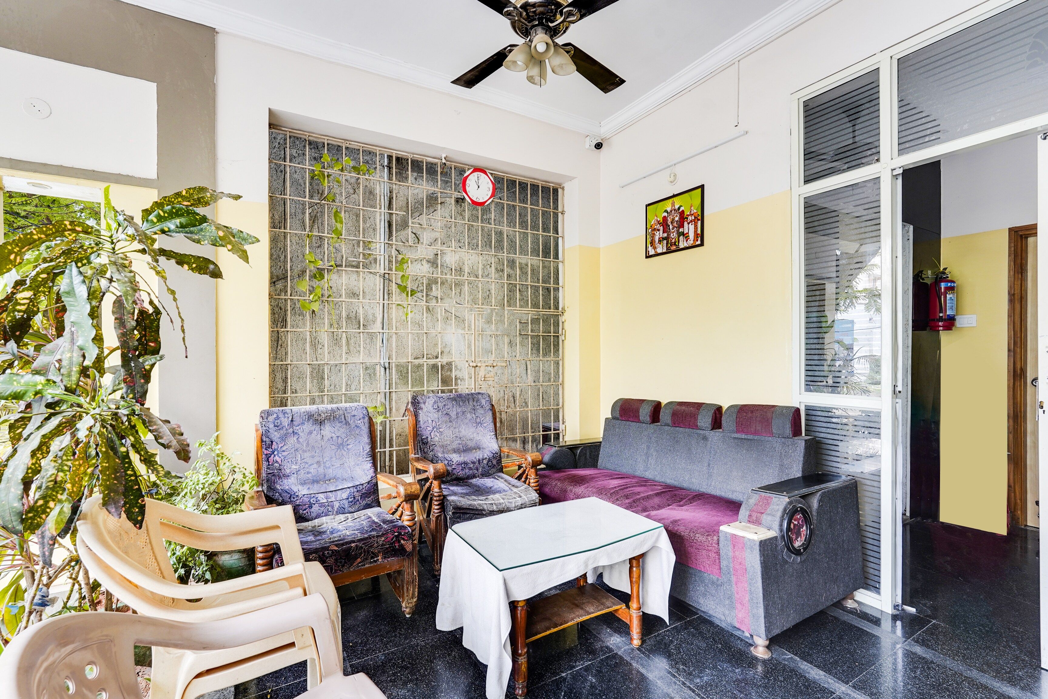 Quarto com sofá, cadeiras, mesa, planta em vaso, janela com grade de metal, ventilador de teto e extintor de incêndio.