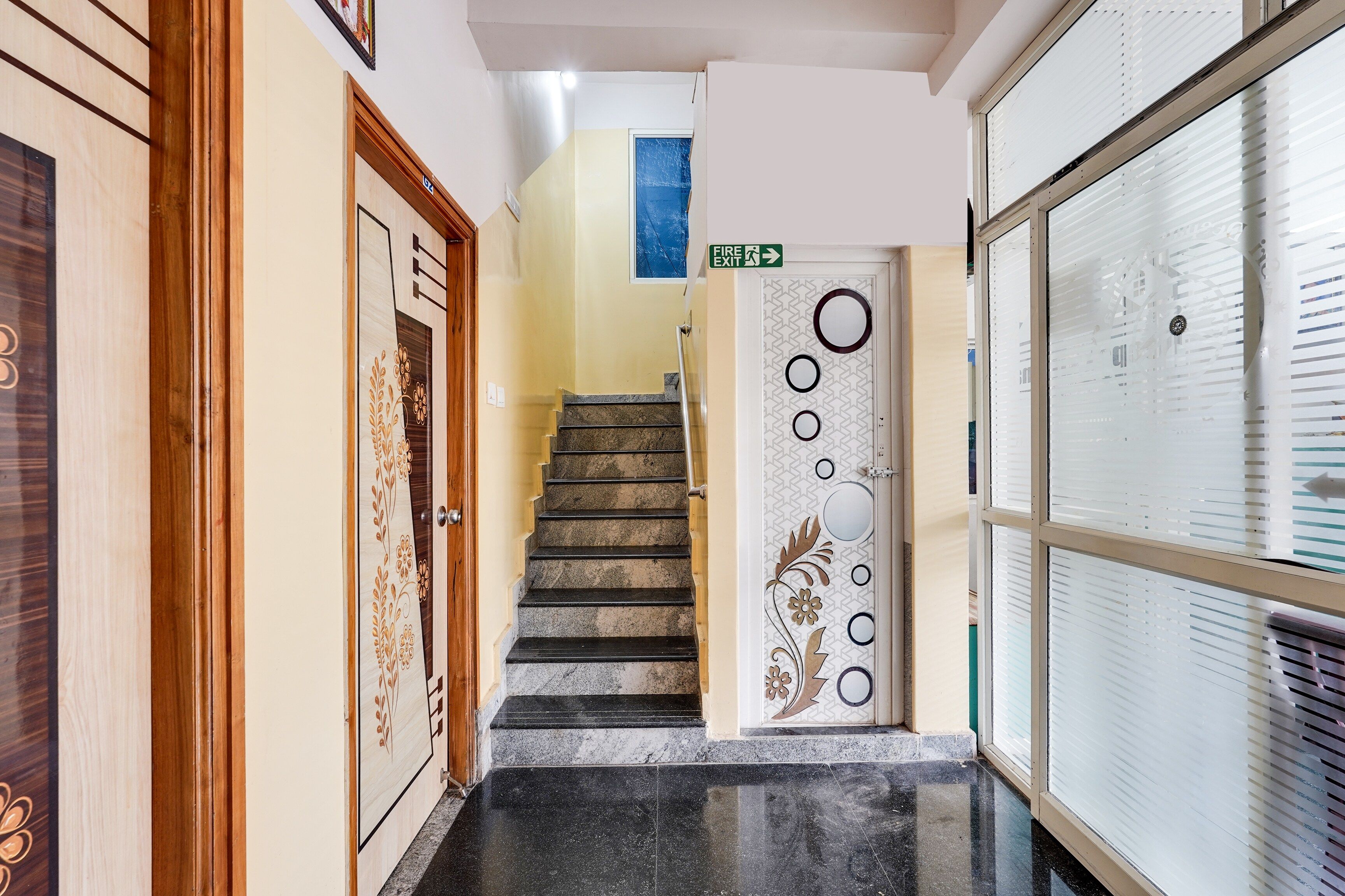 Un passadís amb una porta de fusta a l'esquerra, una escala que mena a una habitació i una porta amb un senyal d'eixida d'incendis a la part superior.
