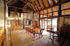 A spacious dining area with a long wooden table and chairs, surrounded by a rustic interior with wooden beams and a thatched roof.
