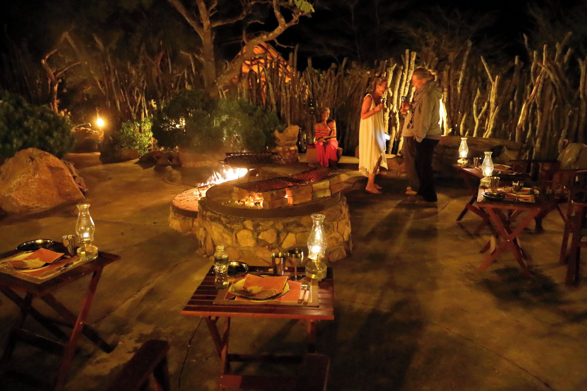 Un espace extérieur aménagé pour les repas, avec des tables et des chaises en pierre, un foyer et un décor rustique agrémenté d’arbres et d’une clôture en bois.