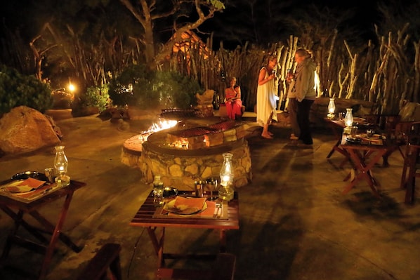 Un espace extérieur aménagé pour les repas, avec des tables et des chaises en pierre, un foyer et un décor rustique agrémenté d’arbres et d’une clôture en bois.