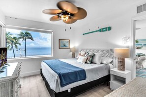 Un dormitorio con cama, ventilador de techo, ventana con vista a palmeras y al océano, y un mapa colgado en la pared.