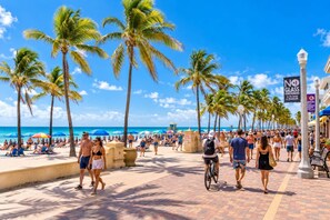 Paseo marítimo con palmeras, gente caminando y un cartel que dice 'NO GLASS CONTAINERS'.