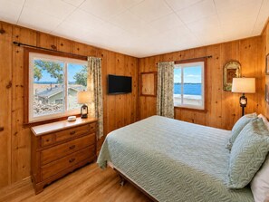 Habitación con paneles de madera, una cama, un aparador, un televisor y ventanas con cortinas.