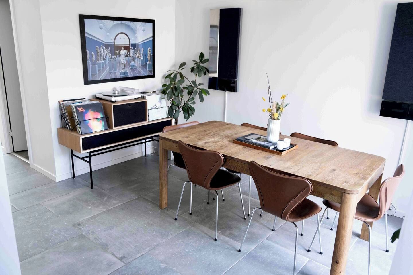 Et spiseområde med et træbord, seks stole, en bænk, en plante, et indrammet billede og en reol med bøger og en vase.