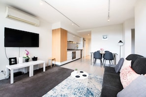 Un salon moderne doté d’un téléviseur à écran plat, d’un banc blanc, d’un coin repas avec une table et des chaises, et d’une cuisine en arrière-plan.