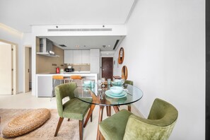 A modern kitchen with white cabinets, a central island, and a dining area with a glass table and green upholstered chairs. - StayShort - Chic 1 Bedroom Haven Perfect for 2 in Dubai Marina! (Dubai Marina)