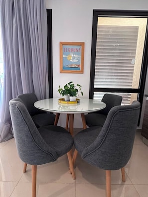 Een eethoek met een ronde glazen tafel, twee grijs gestoffeerde stoelen, een potplant en een ingelijst schilderij aan de muur.