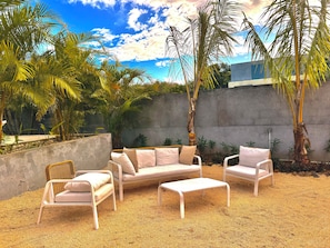 An outdoor seating area with wicker furniture and cushions, surrounded by palm trees and a concrete wall. - The Pearl Villa - With private pool (Petit Raffray)