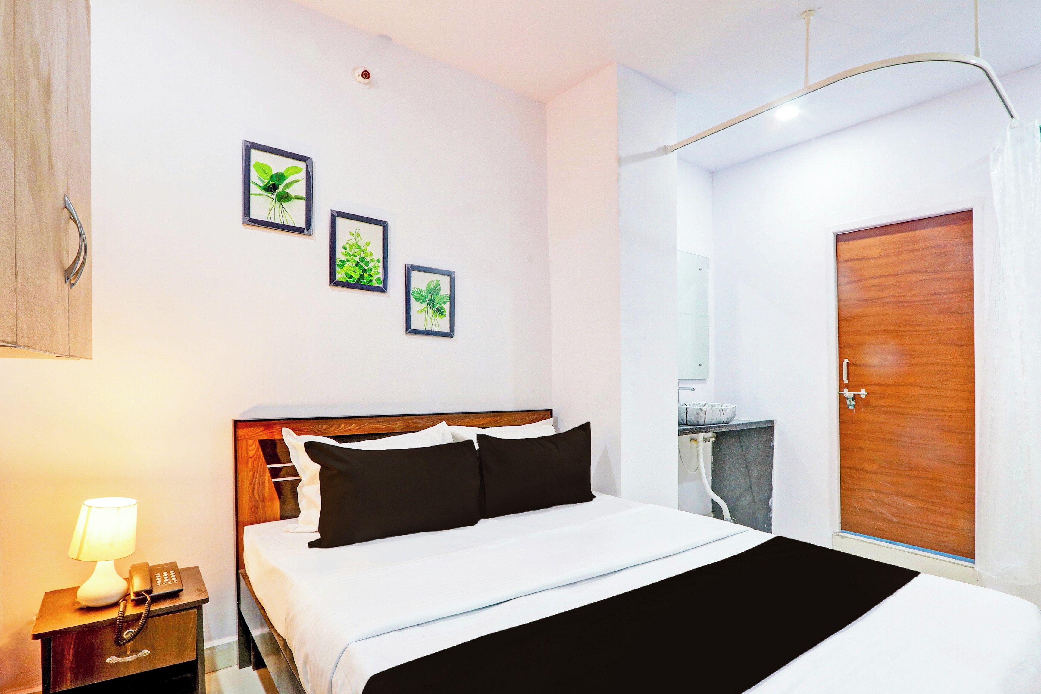 Quarto de hotel com cama, mesa de cabeceira com abajur e telefone, porta de madeira e três quadros de plantas na parede.