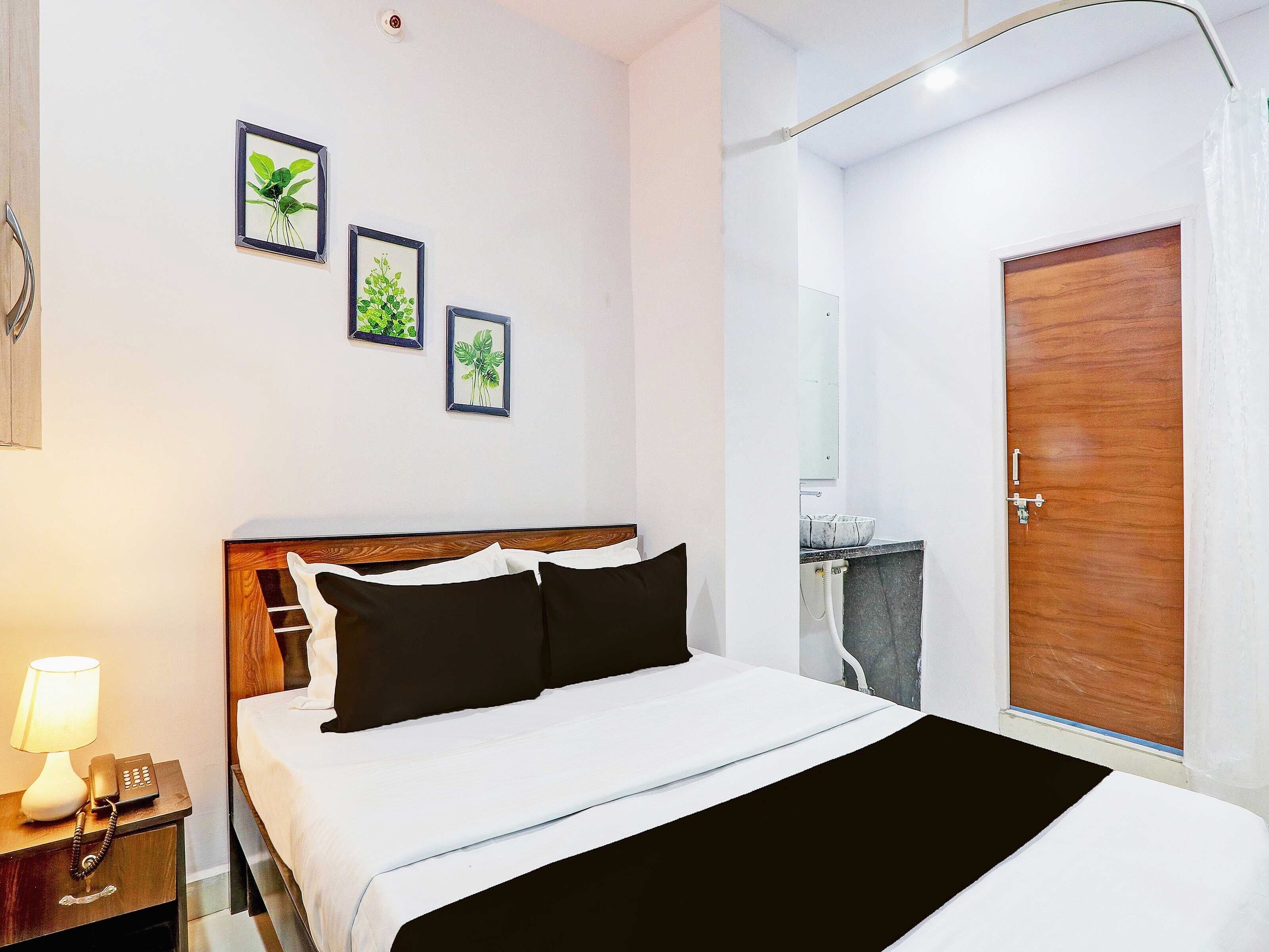 A hotel room with a bed, bedside table with a lamp and phone, a wooden door, and a small bathroom with a sink and mirror.
