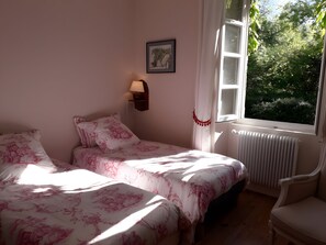 Een slaapkamer met twee bedden, een wandlamp, een radiator en een raam met uitzicht op groen.