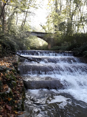 A waterfall cascading over a stone structure with a bridge overhead, surrounded by dense foliage.