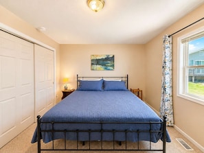Um quarto com cama, mesa de cabeceira, abajur, quadro na parede e janela com cortinas.