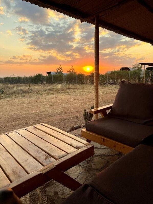 A wooden bench and a cushioned chair under a shelter, with a view of the sunset over a grassy field.
