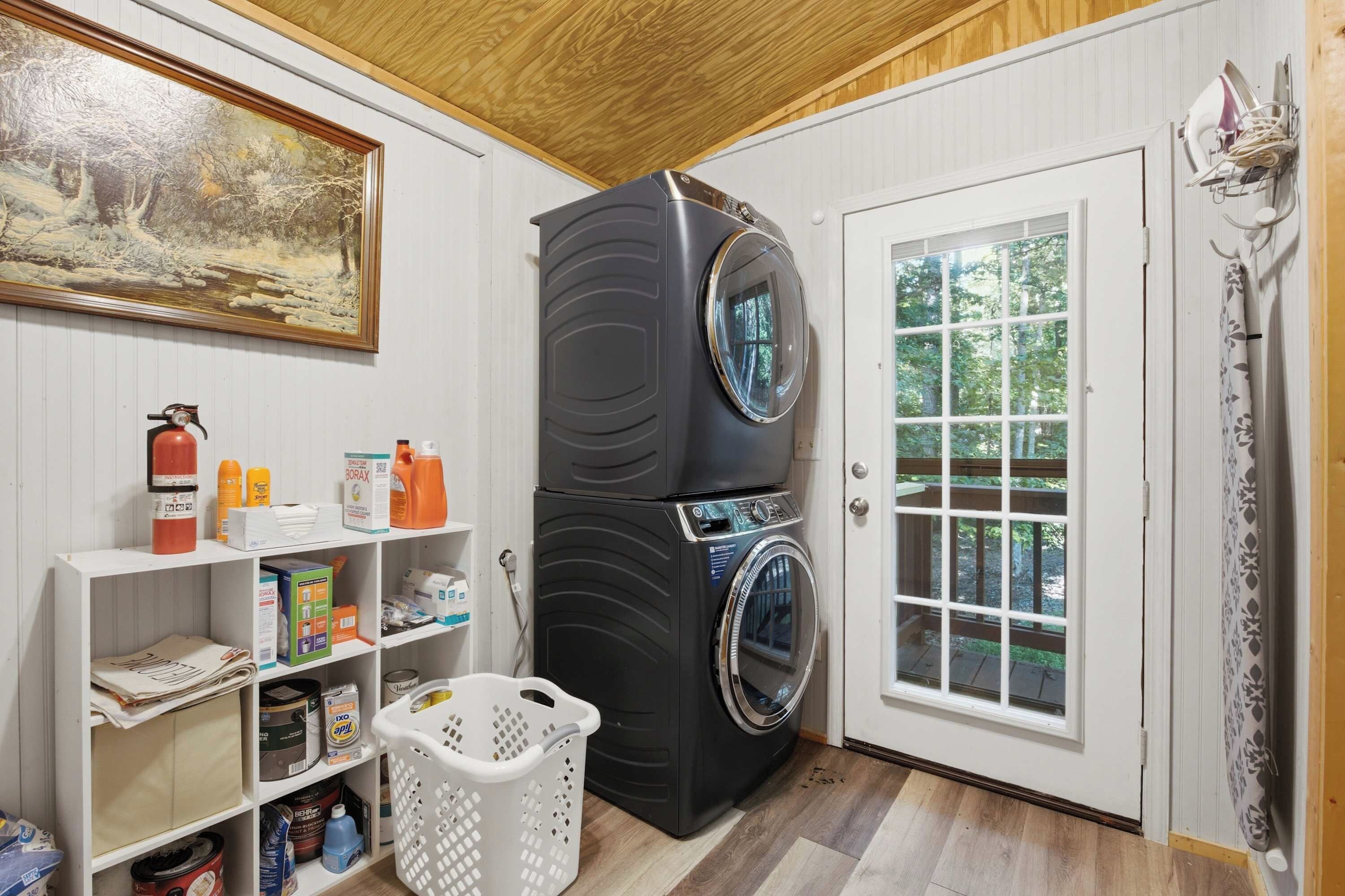 A laundry room with a stacked washer and dryer, a fire extinguisher, cleaning supplies, and a painting on the wall.