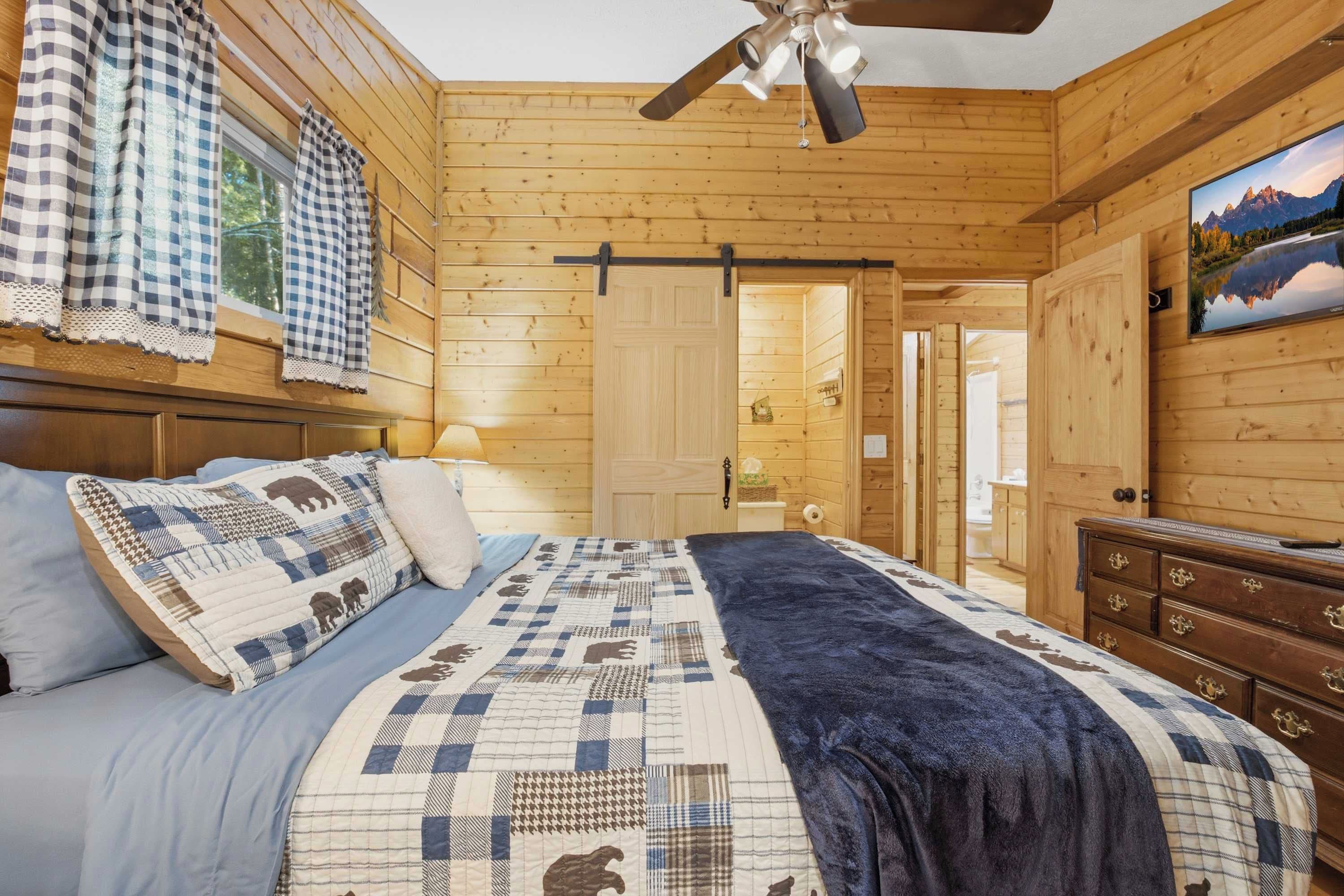 A wooden cabin bedroom with a bed, a ceiling fan, a window with curtains, and a painting on the wall.
