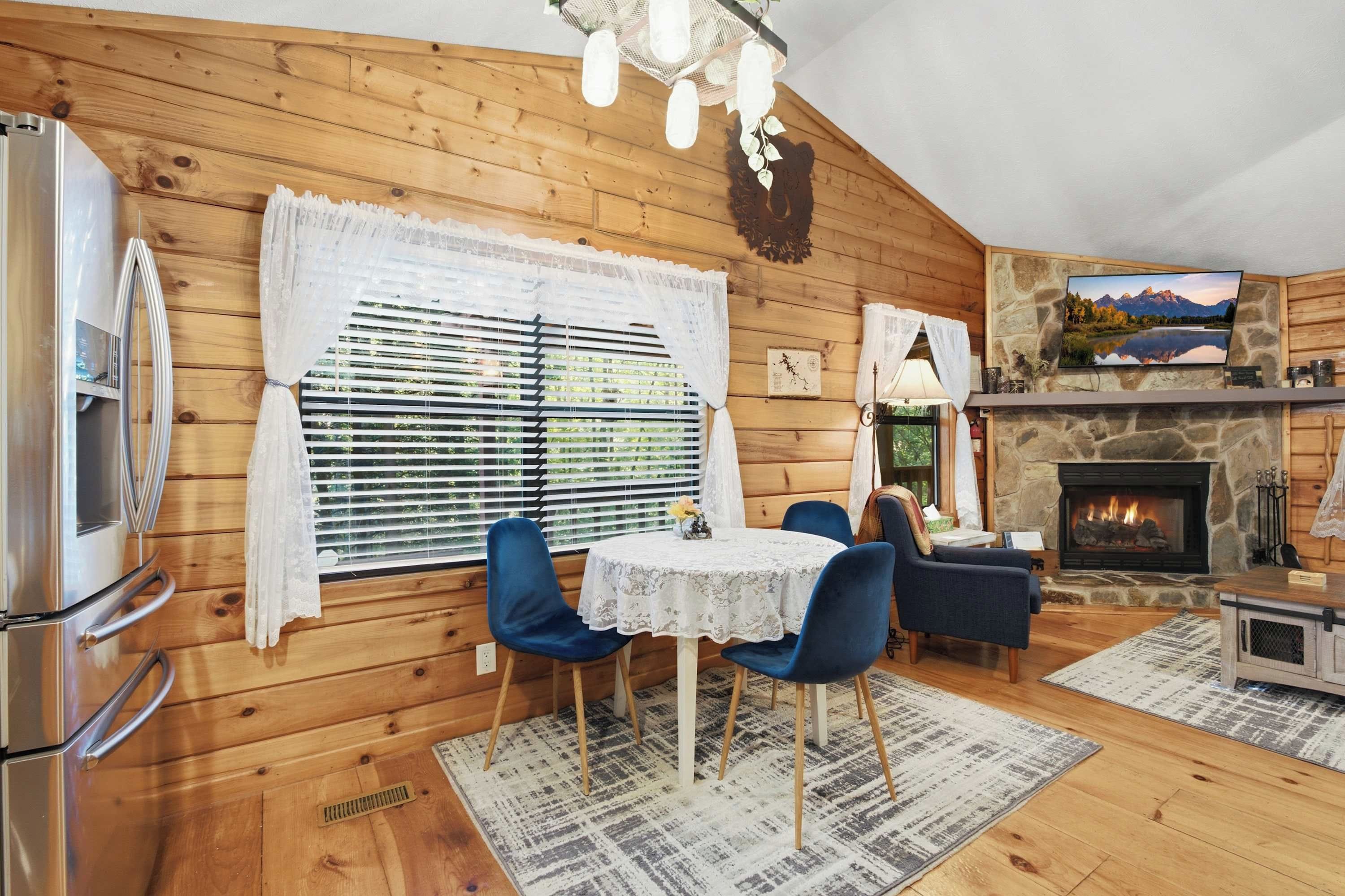 A cozy cabin interior with a stone fireplace, wooden walls, a dining area with blue chairs, and a television mounted above the fireplace.