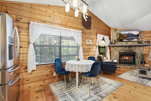 A cozy cabin interior with a stone fireplace, wooden walls, a dining area with blue chairs, and a television mounted above the fireplace. - Bear Essentials & Hot Tub (Blairsville)