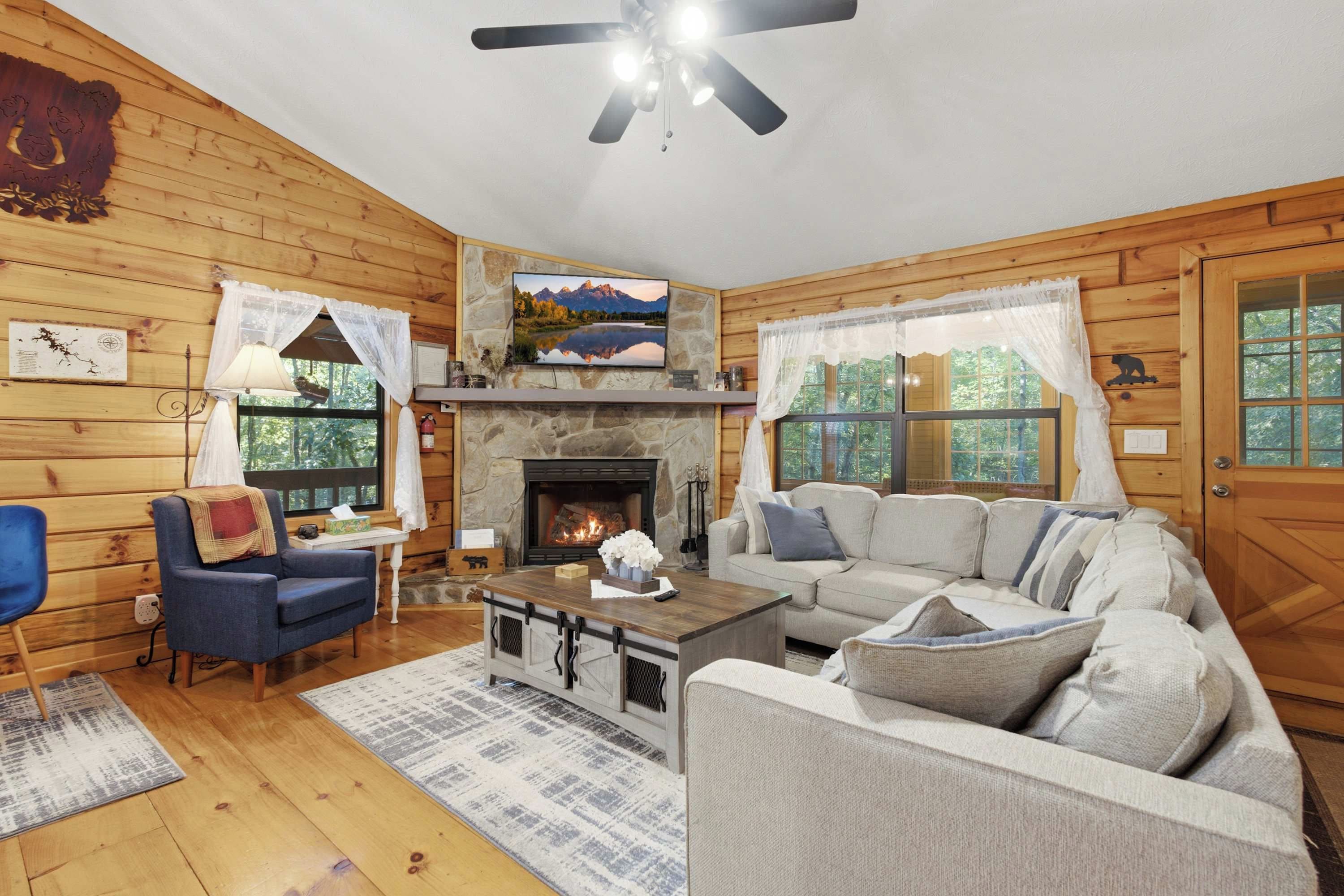 A cozy cabin interior with a fireplace, wooden walls, a sofa, a blue armchair, and a television mounted above the fireplace.