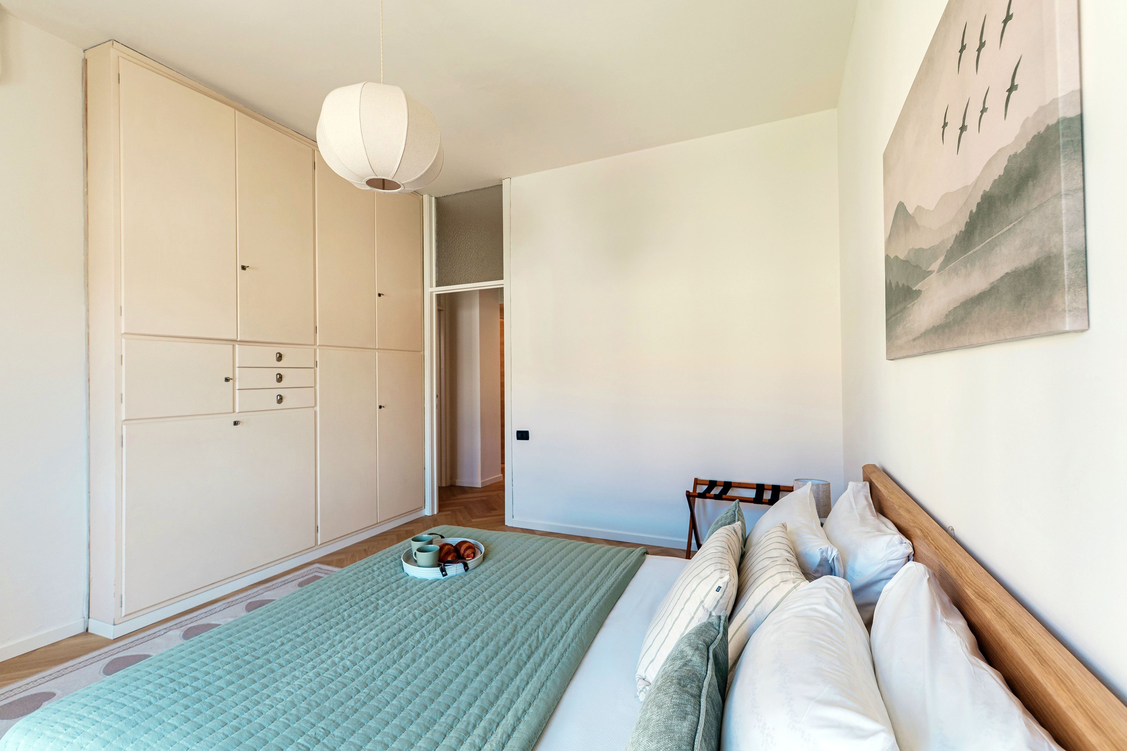 A bedroom with a bed, a wardrobe, a hanging light fixture, and a painting on the wall.